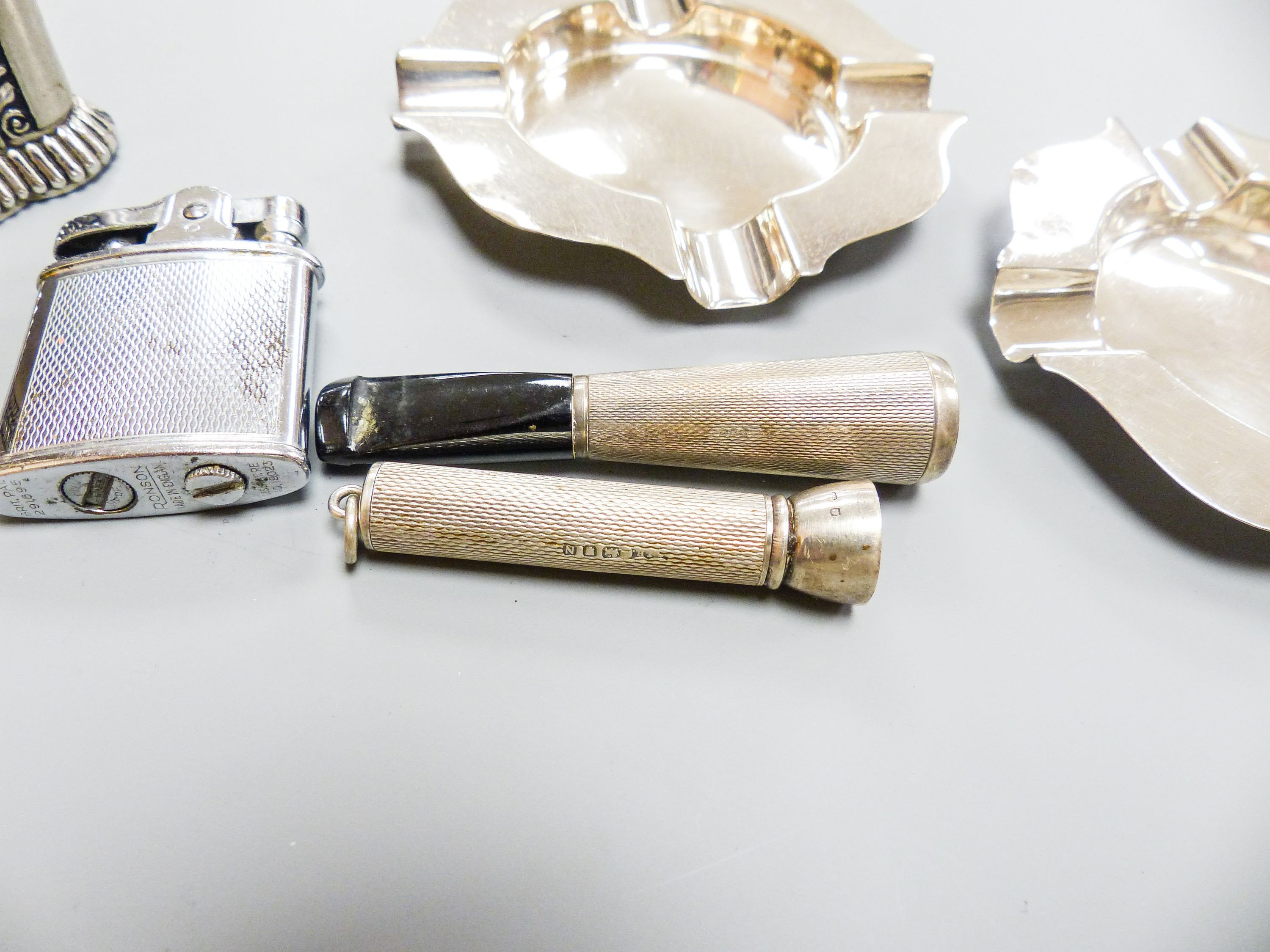 A pair of modern silver ashtrays, two metal lighters, a silver mounted cigar piercer and a white metal cigarette holder.